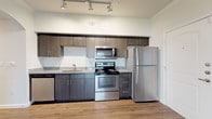 This 1-bedroom, 1-bathroom unit offers a thoughtfully designed single-floor layout that maximizes functionality and comfort. The kitchen is outfitted with stainless steel appliances including a refrigerator, range with microwave above, and dishwasher, all complemented by dark wood-tone cabinetry and granite-style countertops. Track lighting overhead and light wood-look flooring give the kitchen a clean, contemporary feel. An elegant arched doorway connects the kitchen area to the living space, adding a subtle architectural detail that enhances the overall flow of the unit. The living area features crown molding, a ceiling fan with light fixture, and a sliding glass door fitted with vertical blinds that allows natural light to fill the room and provides direct outdoor access. The bedroom is carpeted for comfort and includes a window with horizontal blinds, offering a quiet, restful environment. The bathroom is a standout, featuring granite-style countertops, dark cabinetry, a soaking tub with tile surround, and a stacked in-unit washer and dryer — a highly practical amenity. Throughout the unit, neutral tones and consistent finishes create a cohesive, modern aesthetic.