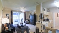This single-floor space showcases an open-concept layout that seamlessly connects the living area and kitchen. The living room is finished with plush carpeting, crown molding, and a ceiling fan with a light fixture that adds both style and functionality. Large sliding glass doors flood the space with natural light while providing direct access to an outdoor patio, offering views of the surrounding exterior. Vertical blinds allow for flexible light control throughout the day. The living area comfortably accommodates a seating arrangement centered around a glass-top coffee table, complemented by a dark wood armoire that serves as a focal point along the back wall. A decorative column with a capital detail elegantly separates the living space from the kitchen area. The kitchen features light-toned cabinetry and a peninsula with bar-height seating, providing additional dining and prep space. The neutral warm color palette throughout—encompassing beige walls and light ceilings—creates a cohesive and inviting ambiance. Decorative accents, including tall candle holders and framed artwork, add personality and refinement to the overall aesthetic.