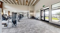 This expansive fitness center offers a generously sized, single-floor layout designed to accommodate a full range of workout activities. The space is divided into distinct functional zones, including a well-equipped strength training area, a cardio section, and a separate open-floor studio ideal for group exercise, stretching, or functional training.

The architectural design blends industrial and contemporary elements, featuring exposed wooden ceiling beams, brick accent columns, and textured tile wall panels that add visual character throughout. Soaring ceilings amplify the sense of openness, while large floor-to-ceiling windows and glass doors flood the interior with natural light and provide views of the surrounding outdoor areas.

The main fitness floor is finished with durable patterned carpet tile and hosts a comprehensive selection of equipment, including cable machines, selectorized strength machines, free weights, dumbbells, and cardio equipment such as treadmills and stationary bikes. Full-length mirrors are strategically placed to support training visibility. The separate studio area features light hardwood flooring, a ceiling fan, wall-mounted displays, and a medicine ball rack, creating an inviting space for dynamic workouts. Pendant lighting and warm accent fixtures contribute to a polished, welcoming ambiance throughout the facility.