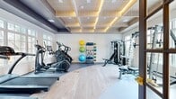 Located at Avalon Haven in North Haven, Connecticut, this well-appointed fitness center offers a thoughtfully designed workout environment across a single open floor. The space is defined by its striking coffered ceiling clad in warm wood paneling with integrated ambient lighting, creating an upscale yet energizing atmosphere. Light hardwood flooring spans the main exercise area, transitioning to durable rubber matting where cardio and strength equipment are stationed.

The cardio zone is generously equipped with treadmills, elliptical trainers, and a stationary bike, all positioned to face large multi-pane windows that frame views of the surrounding greenery, keeping the space bright and connected to the outdoors. On the opposite side, a multi-functional cable and Smith machine system anchors the strength training area, complemented by an adjustable bench and free weight accessories. A centrally located rack displays an array of medicine balls, resistance accessories, and stability balls, supporting diverse functional training routines.

The entry area features textured accent walls with warm cove lighting, a glass-paned interior door, and clean, minimal finishes that maintain a polished, welcoming feel. Ample windows throughout ensure consistent natural light, enhancing the open and airy quality of the space. This fitness center is a well-rounded amenity suited for a full spectrum of workout needs.