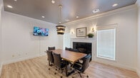 Located at 25455 Borough Park Dr in Spring, TX, this well-appointed conference room offers a sophisticated and functional setting for professional gatherings. The room is finished with crisp white walls, elegant crown molding, and light hardwood flooring that together create a clean, inviting atmosphere. A distinctive black-and-gold tiered pendant light serves as the visual centerpiece, suspended above a stunning live-edge walnut conference table surrounded by eight black leather executive chairs with gold-accented casters. A tile-surround fireplace anchors one wall, framed by a classic white mantel and accented by a framed equestrian artwork and decorative wall sconces, adding warmth and character to the space. Natural light filters in through white-shuttered windows on either side of the fireplace. A wall-mounted flat-screen television is positioned for clear visibility from all seating positions, making it well-suited for presentations or media sharing. Recessed lighting supplements the pendant fixture, ensuring consistent, professional illumination throughout. Abstract wall art adds a modern touch, while the overall design strikes a balance between corporate functionality and refined, comfortable aesthetics — making this conference room a compelling venue for team meetings, client consultations, and collaborative sessions.