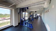 Located at 1304 Big Bend Rd in Twin Oaks, Missouri, this single-floor fitness center is a well-appointed facility designed to support a comprehensive workout experience. The space features an open, industrial-style aesthetic with exposed structural ceilings, painted ductwork, and linear LED lighting that illuminates the entire facility efficiently.

The main workout floor is lined with large windows that flood the space with natural light and offer views of the surrounding area. It is equipped with a robust selection of cardio machines — including treadmills and ellipticals — alongside blue-framed cable machines, weight benches, barbells, and a full dumbbell rack. Durable rubber flooring throughout the exercise areas provides safety and support during training.

A dedicated studio room, enclosed by full-height glass partition walls with black frames, offers space for group fitness classes or cycling, with smooth hardwood-style flooring suited for varied activities. Adjacent to the studio is a lounge corridor with small bistro tables and chairs, providing a space to rest or gather.

The facility also includes a welcoming entrance corridor with posted fitness center rules, restroom access, and a water fountain. The thoughtful layout ensures a smooth flow between zones, making this fitness center a functional and modern destination for residents or members seeking a complete exercise environment.