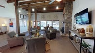 This single-story space is defined by its warm, rustic character and thoughtfully designed open layout. Exposed wooden beams span the vaulted ceiling overhead, adding architectural depth and a sense of craftsmanship that sets the tone throughout the interior. Pendant lighting hangs from the beams, casting a welcoming glow across the space.

At the heart of the room, a floor-to-ceiling stacked stone fireplace serves as a commanding focal point, anchoring the lounge area with natural texture and visual drama. The seating area is furnished with plush, neutral-toned armchairs arranged around a central coffee table, creating an inviting space for relaxed conversation.

Beyond the lounge, a dining area is positioned near large windows that allow generous natural light to filter in, offering views of the surrounding outdoor landscape. The transition between the two areas flows naturally, supported by the open plan and consistent warm, earthy color palette used throughout.

Decorative wall art and carefully selected greenery add finishing touches that enhance the welcoming atmosphere. The combination of natural materials — stone, wood, and woven textiles — gives the space a grounded, cohesive aesthetic that balances comfort with character. Overall, the space presents a well-appointed, community-oriented environment with a distinctive lodge-inspired design sensibility.
