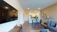This single-floor residence offers a thoughtfully designed open-concept layout that effortlessly connects the living, dining, and kitchen areas into one cohesive and functional space. The living area anchors the layout with a wall-mounted flat-screen television and tasteful décor accents, creating a welcoming atmosphere for relaxation and entertainment. Wide-plank hardwood-style flooring flows throughout, lending warmth and visual continuity to the entire space. The dining area features a round glass-top table with sleek chairs, positioned to allow easy interaction with both the living and kitchen areas. The kitchen is equipped with stainless steel appliances, including a full-size refrigerator, white shaker-style cabinetry, and a peninsula that serves as both a workspace and a casual seating option. Recessed lighting throughout ensures bright, even illumination across the open floor plan. The clean, neutral color palette—featuring crisp white ceilings and soft warm-toned accent walls—contributes to an airy, modern ambiance. The space also includes interior doorways leading to additional rooms, providing a well-organized and practical flow. Overall, the design balances contemporary style with everyday functionality.