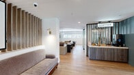 Located at 2850 Kalmia Ave in Boulder, Colorado, this single-floor space offers a generous and well-appointed layout designed for both collaboration and comfort. The open-plan kitchen and living room serve as the heart of the space, showcasing light wood flooring throughout that lends warmth and continuity to the design. The kitchen is a standout feature, boasting dual white quartz countertops — including a large central island — flanked by sleek grey-blue cabinetry with brushed gold hardware. A series of pendant lights hang above, while a full-length window at the far end floods the space with natural light. Adjacent to the kitchen, a dedicated coffee bar station with dark wood cabinetry and slatted dividers adds a refined, hospitality-inspired touch. The living area is divided into distinct lounge zones, each thoughtfully furnished with contemporary seating, large-scale artwork, and statement pendant lighting. A tray ceiling detail crowns the main lounge, adding architectural depth. A glass-enclosed private room with bold black framing provides a quiet retreat. The hallway connects the zones seamlessly, with decorative wood slat paneling and brass wall sconces adding texture and elegance throughout.