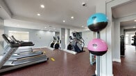 This fitness center is a comprehensively outfitted, single-floor facility designed to support a full range of workout routines. The space is organized into distinct zones, allowing for efficient use of both cardio and strength training equipment throughout the open layout. Structural columns punctuate the floor plan, and large windows along one wall allow natural light to fill the cardio area, where a row of treadmills faces the outdoors. The ceiling features a tray design with recessed lighting throughout, contributing to a clean, well-lit environment.

The cardio section is equipped with treadmills, elliptical trainers, stair climbers, and a Matrix cross-trainer. The strength training area includes multiple selectorized cable machines, a multi-functional power rack with an adjustable bench, and a dedicated free weights section with a full dumbbell rack and stability balls in various sizes. Wall-mounted mirrors in the strength zones enhance visibility and expand the sense of space.

Underfoot, durable speckled rubber flooring covers the entire facility, providing both safety and noise reduction. Practical amenities include a dual-basin water fountain with a bottle-filling station and a mounted fire extinguisher. The walls are painted in a cool grey palette, lending the space a modern, professional atmosphere suited for serious training.