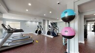 This fitness center is a comprehensively outfitted, single-floor facility designed to support a full range of workout routines. The space is organized into distinct zones, allowing for efficient use of both cardio and strength training equipment throughout the open layout. Structural columns punctuate the floor plan, and large windows along one wall allow natural light to fill the cardio area, where a row of treadmills faces the outdoors. The ceiling features a tray design with recessed lighting throughout, contributing to a clean, well-lit environment.

The cardio section is equipped with treadmills, elliptical trainers, stair climbers, and a Matrix cross-trainer. The strength training area includes multiple selectorized cable machines, a multi-functional power rack with an adjustable bench, and a dedicated free weights section with a full dumbbell rack and stability balls in various sizes. Wall-mounted mirrors in the strength zones enhance visibility and expand the sense of space.

Underfoot, durable speckled rubber flooring covers the entire facility, providing both safety and noise reduction. Practical amenities include a dual-basin water fountain with a bottle-filling station and a mounted fire extinguisher. The walls are painted in a cool grey palette, lending the space a modern, professional atmosphere suited for serious training.