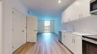 This contemporary single-floor unit presents a clean, modern aesthetic throughout, defined by crisp white walls, wood-look luxury vinyl plank flooring, and an abundance of natural light. The open-concept living and kitchen area forms the heart of the unit, where a streamlined kitchen features white flat-panel cabinetry with brushed nickel hardware, a white penny-tile backsplash, a stainless steel sink, and a full suite of stainless steel appliances including a refrigerator, electric range, over-the-range microwave, and dishwasher. The living area flows seamlessly from the kitchen, with a large window framing views of the surrounding neighborhood and filling the space with natural light. A dedicated bedroom offers a quiet retreat, highlighted by a tall, narrow window that draws in daylight while maintaining privacy and providing outdoor views. The unit includes the convenience of in-unit stacked washer and dryer, neatly housed in a dedicated closet. The bathroom is finished with large-format neutral tile flooring, white subway tile surrounding a full soaking tub, and a clean, minimalist vanity. Throughout the unit, the consistent use of light tones and modern materials creates a bright, cohesive, and highly functional living environment.