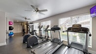 Located at 4010 Linkwood Dr in Houston, Texas, this fitness center offers a comprehensive workout facility spread across a single floor. The space is thoughtfully designed to accommodate a wide range of exercise needs, with distinct zones for cardio training, strength work, and functional fitness. Large windows fitted with horizontal blinds line the walls, flooding the interior with natural light while offering views of the surrounding neighborhood. Ceiling fans are positioned throughout the facility, ensuring comfortable airflow during workouts. The durable rubber-textured flooring provides both safety and practicality across the exercise areas, while a section of hardwood flooring near the entry adds a welcoming touch. The cardio zone features multiple treadmills, elliptical trainers, and a stair stepper, while the strength area is anchored by a multi-station cable machine and a dedicated free weight bench. A stretching and functional fitness corner is stocked with stability balls and a gymnastics bar, rounding out the offerings. A wall-mounted display screen and posted fitness information contribute to an organized, user-friendly environment. The space is accessible via a dedicated entrance door, making it a convenient and well-appointed fitness destination.