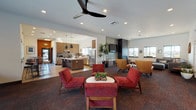 Located at 760 Central Avenue in Moses Lake, Washington, this single-story community clubhouse delivers a well-appointed, contemporary gathering space designed for both socializing and productivity. The entrance welcomes visitors with light wood-look flooring that transitions into richly patterned carpet throughout the main areas.

The open-plan lounge and kitchen area serves as the heart of the space. A full community kitchen features warm walnut cabinetry, quartz countertops, a central island with pendant lighting, a mosaic tile backsplash, and stainless steel appliances. Adjacent bar-height seating along the windows provides a casual dining option with natural light.

The lounge area is divided into distinct seating zones, including a conversation area with bold red accent chairs, a relaxed sectional sofa nook with a decorative wood panel feature wall, and a media area anchored by a sleek floor-to-ceiling black fireplace surround with a wall-mounted flat-screen TV.

A dedicated conference area features two private meeting rooms adorned with retro-patterned wallpaper and large windows overlooking the outdoor courtyard and pool. The leasing or reception area includes a quartz-topped island counter beneath a statement geometric chandelier, with additional offices accessible through dark-framed doors. Throughout, recessed lighting and clean white walls create a bright, modern ambiance.