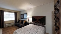 Located at 5350 Silverthorne Road in Westerville, Ohio, this single-floor room offers a well-appointed and comfortable retreat. The space is designed with functionality and relaxation in mind, featuring a queen-sized bed dressed in patterned bedding and accented by a bold geometric headboard in rich mahogany and black tones. A nightstand with a classic table lamp provides warm, ambient lighting, complementing the neutral wall tones throughout. A cozy seating area with a dark upholstered sofa and decorative throw pillows offers a comfortable spot to unwind, making the room suitable for both rest and leisure. Two framed art pieces adorn the wall above the sofa, adding a polished, curated touch to the décor. A window with dark curtains allows for light control and privacy. The textured ceiling and light wood-toned flooring contribute to the room's understated, welcoming aesthetic. The overall ambiance is calm and practical, with a layout that makes efficient use of the available space while maintaining a comfortable and inviting atmosphere for occupants.