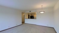 This single-floor space features an open-concept design that connects the living area to the kitchen through a half-wall pass-through, creating a sense of openness and visual continuity. The living area is finished with wall-to-wall carpeting in a neutral tone, complemented by clean white walls and dark baseboards that frame the space neatly. A pendant light fixture with a bowl-shaped shade hangs overhead, providing a focal point and ambient lighting to the main living space. The kitchen, visible through the pass-through opening, is equipped with light wood cabinetry and a black refrigerator. The countertop along the pass-through features a granite or stone-style surface, adding a touch of character. A HVAC vent is positioned in the ceiling near the kitchen opening, ensuring climate control throughout. The layout allows for a natural flow between the living and kitchen areas, making the space functional and easy to navigate. A hallway visible to the left suggests additional rooms beyond, contributing to the overall practical layout of this well-maintained, move-in ready space.