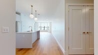 Located at 695 Merrick Ave in Westbury, NY, this single-floor residence showcases a clean, contemporary design with high-quality finishes throughout. The open-concept layout centers around a striking kitchen featuring a large island with a white quartz countertop, a brushed gold faucet, and an undermount sink. Flat-front white cabinetry extends floor to ceiling, complemented by stainless steel appliances including a French door refrigerator, gas range, and built-in microwave. Globe pendant lights on brass fixtures hang above the island, adding a warm, modern touch. Wide-plank light oak hardwood flooring flows seamlessly from the kitchen through the expansive living area, which is brightened by large windows overlooking the surrounding neighborhood. The bedroom offers generous proportions, its own large window with exterior views, and direct access to a hallway with multiple closets leading to a well-appointed bathroom. A half bath features elegant marble-look tile flooring and polished brass hardware, maintaining the cohesive design language found throughout. Crisp white walls, recessed lighting, and gold-toned accents unify the space with a refined, modern aesthetic.