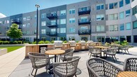 Located at 4500 W Touhy Ave in Lincolnwood, Illinois, this thoughtfully designed outdoor amenity courtyard is nestled within a modern multi-story residential building, offering residents a refined and functional open-air gathering space. The courtyard is framed on multiple sides by the building's contemporary façade, characterized by clean lines, gray horizontal cladding, large windows, and private balconies above.

The outdoor space is anchored by a well-equipped grilling station featuring multiple built-in gas grills set within warm wood-accented cabinetry, ideal for communal cooking and entertaining. Nearby, woven rattan dining chairs paired with teak-topped round tables are arranged throughout the spacious poured-concrete patio, providing ample seating for residents to dine and socialize.

A standout feature is the covered pergola area, which houses a built-in outdoor bar topped with a wall-mounted television and framed by warm wood paneling. Bar-height seating lines the counter, creating an inviting spot for casual gatherings or watching the game outdoors.

Lush ornamental grasses, manicured green lawn sections, and strategically placed trees soften the hardscape and add a natural, park-like quality to the environment. The courtyard's open layout maximizes natural sunlight, making it a bright and welcoming retreat throughout the day.