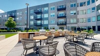 Located at 4500 W Touhy Ave in Lincolnwood, Illinois, this thoughtfully designed outdoor amenity courtyard is nestled within a modern multi-story residential building, offering residents a refined and functional open-air gathering space. The courtyard is framed on multiple sides by the building's contemporary façade, characterized by clean lines, gray horizontal cladding, large windows, and private balconies above.

The outdoor space is anchored by a well-equipped grilling station featuring multiple built-in gas grills set within warm wood-accented cabinetry, ideal for communal cooking and entertaining. Nearby, woven rattan dining chairs paired with teak-topped round tables are arranged throughout the spacious poured-concrete patio, providing ample seating for residents to dine and socialize.

A standout feature is the covered pergola area, which houses a built-in outdoor bar topped with a wall-mounted television and framed by warm wood paneling. Bar-height seating lines the counter, creating an inviting spot for casual gatherings or watching the game outdoors.

Lush ornamental grasses, manicured green lawn sections, and strategically placed trees soften the hardscape and add a natural, park-like quality to the environment. The courtyard's open layout maximizes natural sunlight, making it a bright and welcoming retreat throughout the day.