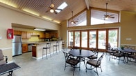 This single-story clubhouse is designed to serve as a welcoming community gathering space, combining functional amenities with an inviting architectural character. The main gathering area immediately draws attention with its dramatic vaulted ceiling clad in warm wood paneling, accented by ceiling fans and skylights that fill the space with natural light. A striking floor-to-ceiling stone fireplace anchors one end of the room, providing a rustic focal point surrounded by comfortable seating arranged for conversation.

Large wood-framed French doors line one wall, offering direct views and access to an outdoor pool area, seamlessly connecting the interior to the exterior amenity space. Tile flooring runs throughout, and iron-framed dining sets provide casual seating across multiple areas of the main room.

The full kitchen is well-equipped with warm oak cabinetry, stainless steel appliances including a range, dishwasher, and refrigerator, and generous counter space. A peninsula counter with bar seating extends into the main area, making it practical for serving during events.

Additional features include two restrooms with accessible-height vanities and tile flooring, a cozy lounge nook near the fireplace, and decorative artwork displayed throughout, contributing to the clubhouse's warm and functional ambiance.