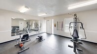 Located at 1840 Killingsworth Rd in Augusta, Georgia, this dedicated exercise room offers a practical and functional fitness facility within a compact, single-floor layout. The space features durable rubber flooring throughout, providing a safe and resilient surface ideal for a variety of workout activities. Bright fluorescent lighting illuminates the room evenly, complemented by natural light from multiple windows fitted with blinds. A textured ceiling adds a classic architectural touch to the interior.

The exercise room is outfitted with a well-rounded selection of fitness equipment, including stationary bikes, an elliptical machine, and a multi-function cable weight machine, catering to both cardio and strength training needs. Colorful resistance bands are mounted neatly along one wall for added versatility. A large wall-mounted mirror spans a significant portion of one wall, visually expanding the space and allowing users to monitor their form during workouts.

A centrally placed utility counter with open shelving provides convenient storage for accessories such as yoga mats, resistance tools, and small equipment. A compact mini-fridge is integrated beneath the counter for added convenience. The room also includes access to restroom facilities and is climate-controlled via a wall-mounted air conditioning unit, ensuring year-round comfort for users.