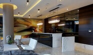 This single-floor space presents a striking contemporary lobby designed with meticulous attention to detail and high-end finishes. The expansive marble flooring, rendered in cool grey and white tones with natural veining, establishes an immediate sense of refinement and grandeur throughout the space.

A prominent cylindrical column anchors the center of the lobby, finished with a warm gold-toned ceiling detail that introduces a touch of opulence against the otherwise neutral palette. Recessed cove lighting runs along the ceiling perimeter, casting a soft, ambient glow that complements the cluster pendant and chandelier-style fixtures suspended above key areas.

The reception area features a sleek marble-fronted counter paired with a dark, glossy back panel that creates a bold visual contrast. A dramatic abstract wall mural in warm amber and gold tones serves as a commanding focal point behind the desk.

The seating area includes modern white leather chairs arranged around a low central table, accented by a floral arrangement and set atop a sculptural area rug. Large windows along one side introduce natural light, enhancing the bright, open ambiance of this elegantly appointed space.