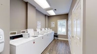 Located at 2605 Elms Plantation Blvd in Charleston, South Carolina, this shared laundry facility offers a practical and well-maintained space designed for convenient communal use. The single-floor layout is efficiently organized, featuring a row of top-loading washing machines aligned along one wall, complemented by a bank of dryers along the opposite side, providing ample capacity for residents. The room is finished with warm taupe-toned walls and durable wood-look vinyl plank flooring, lending a clean and modern aesthetic. Overhead fluorescent lighting ensures the space is bright and functional at any time of day. A window with blinds allows natural light to filter in, enhancing the overall ambiance. Instructional signage for both washing and drying is posted on the walls, offering helpful guidance for users. A waste bin is conveniently placed within the room to support cleanliness and upkeep. The space is thoughtfully designed to accommodate multiple users simultaneously, making it a highly practical shared amenity. The overall atmosphere is tidy, organized, and welcoming, reflecting a commitment to providing a quality laundry experience for the community it serves.