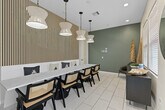 Located at Arrive at Grand Terrace in Moreno Valley, California, this thoughtfully designed community room offers a refined and welcoming environment. The space is anchored by a striking vertical slatted wood accent wall, which serves as a dramatic backdrop for a long, white marble-topped communal table surrounded by elegant cane-back chairs with black frames — an ideal setting for meetings, gatherings, or collaborative work. A row of sculptural white pendant lights hang in sequence above the table, casting warm, even illumination throughout the room. The light tile flooring adds brightness and a clean, contemporary feel underfoot. Sage green walls create a calm, sophisticated atmosphere that pairs seamlessly with the natural textures of the wood slat feature and decorative dried pampas grass arrangements. A glass entry door allows natural light to filter in and provides a view of the outdoor surroundings. Along one wall, a sleek dark credenza with glass-front cabinetry holds curated décor accents, while a circular mirror and framed wall art contribute to the polished aesthetic. A leather accent chair offers additional seating. The overall ambiance is modern, warm, and inviting — designed to foster both functionality and style.