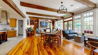 This single-story space blends rustic charm with modern comfort through a thoughtfully designed open layout. Reclaimed wood ceiling beams run throughout the main gathering area, complementing the rich, wide-plank hardwood floors that anchor the space with warmth and character. A prominent brick fireplace serves as a focal point in the living area, flanked by built-in wood shelving with warm amber-toned cabinetry, providing both display and storage. A wall-mounted television above the fireplace mantel adds a contemporary touch. The seating area features generous leather sofas and upholstered armchairs arranged around patterned area rugs, creating an inviting and layered atmosphere. A central dining table with industrial-style metal legs offers a transitional gathering spot between the living and kitchen zones. Floor-to-ceiling windows and French doors flood the interior with abundant natural light and provide direct views of an outdoor patio and pool area. The kitchen is appointed with warm wood cabinetry, white quartz countertops, stone tile flooring, a professional-grade stainless steel range with a hood, a large center island with bar seating, and industrial pendant lighting. The overall ambiance is cohesive, comfortable, and richly detailed.