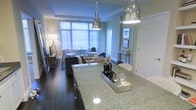 This well-appointed single-floor residence offers a thoughtfully designed open-concept layout that seamlessly connects the kitchen and living areas. The kitchen is a standout feature, showcasing a large granite-topped island with under-counter lighting, dark cabinetry with polished hardware, and a pair of sleek industrial-style pendant lights that hang overhead, adding a contemporary touch. The open flow leads into a comfortable living area anchored by a plush sectional sofa and a warm wooden coffee table, creating an inviting space for relaxation and entertaining. Large windows dressed with horizontal blinds allow generous amounts of natural light to fill the interior, lending the space an airy, open feel. A console table styled with decorative greenery and framed wall art adds a refined accent near the transition between spaces. The flooring transitions from rich dark hardwood in the kitchen and living area to lighter tones, contributing to a dynamic yet cohesive aesthetic throughout. The overall ambiance is modern and sophisticated, with a careful balance of warm and cool tones that creates an elegant and livable environment.