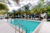 Located at 1361 S Federal Hwy in Boca Raton, Florida, this multi-story property showcases a distinctive white stucco exterior with a clean, contemporary architectural design accented by private balconies on each level. The building spans two floors and is centered around a beautifully curated outdoor amenity area that serves as the focal point of the community.

The expansive resort-style pool is the standout feature, offering crystal-clear turquoise water surrounded by wide tiled decking lined with rows of modern chaise lounges and navy blue market umbrellas. Towering mature palm trees frame the pool on all sides, lending a tropical ambiance that is quintessentially South Florida.

Beyond the pool, lush green lawn areas are meticulously landscaped with tropical foliage, decorative river rock beds, and manicured hedgerows. Wide paved pathways connect the various outdoor zones, including relaxation areas furnished with woven lounge chairs, hanging swing seats, and hammocks nestled among the palms. A cornhole game court and additional seating vignettes provide recreational options throughout the grounds.

The building's facade, visible from the courtyard, features iron-railed balconies overlooking the verdant setting, creating a seamless indoor-outdoor connection. Rich tropical plantings — including crotons, ficus, bamboo, and palms — are thoughtfully arranged throughout, enhancing both privacy and visual appeal across the entire property.