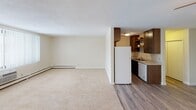 This single-floor residential unit offers a well-organized layout designed for comfortable everyday living. The open living area is generously proportioned, featuring soft wall-to-wall carpeting, a textured ceiling, and baseboard heating that runs along the perimeter. Vertical blinds on the windows provide flexible light control, allowing natural light to filter through and brighten the space. The living area flows directly into the kitchen, which transitions to durable hardwood-style flooring. The kitchen is outfitted with dark wood cabinetry, open shelving, a white refrigerator, a dishwasher, and a sink with a window view — offering a practical and efficient workspace. Two bedrooms provide comfortable retreats, both finished with plush carpeting and baseboard heating. One bedroom features a ceiling fan with a light fixture, while the other includes a walk-in closet with a hanging rod, complemented by an additional closet for extra storage. The full bathroom is cleanly finished with white tile, a tub-shower combination, a wide vanity with a single sink, a large mirror, and a stylish three-bulb light fixture above. Throughout, the neutral color palette and consistent finishes create a cohesive, move-in-ready environment.