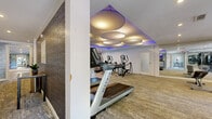 Located at 1500 Harbor Landing in Roswell, Georgia, this well-appointed fitness facility spans a generous single floor and is divided into two distinct functional areas designed to support a comprehensive workout experience. The main fitness floor is a spacious, open exercise room outfitted with a full lineup of Matrix cardio equipment, including multiple treadmills and elliptical trainers, as well as rowing machines, cable strength systems, and a free weight area with dumbbells and benches. Floor-to-ceiling mirrors line the walls, amplifying the sense of space and allowing members to monitor their form. The room is elevated by a distinctive dropped ceiling with oversized circular pendant light fixtures accented by soft purple ambient lighting, creating a modern, energetic atmosphere. A large-scale typographic wall mural reinforces the fitness theme, and a wall-mounted flat-screen TV keeps members engaged during cardio sessions. Carpeted flooring throughout the main area adds comfort underfoot. The second space serves as a dedicated yoga and stretching studio, featuring warm hardwood-style flooring, full-length mirrors, and large windows that invite natural light and views of the surrounding outdoor area. Built-in shelving stores yoga mats and accessories, while a vibrant nature-inspired wall mural enhances the calm, focused ambiance ideal for stretching and mindfulness practice.