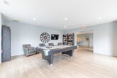 Located at 472 Brock Avenue in Burlington, Ontario, this well-appointed amenity space spans a single floor and is designed to foster community and relaxation. The open-concept layout is anchored by light wood-look flooring and contemporary finishes throughout, creating a cohesive and inviting atmosphere.

The main lounge and dining area serves as the heart of the space, featuring a round marble-top dining table, upholstered seating, and a wall-mounted flat-screen TV. Textured grey accent walls add depth and sophistication, while large windows and a sliding glass door to a balcony flood the area with natural light and offer views of the surrounding Burlington streetscape.

Adjacent to the lounge is a fully equipped kitchen, outfitted with white cabinetry, a subway tile backsplash, full appliances, and a central island with bar stool seating — ideal for hosting and social gatherings. A striking piece of artwork adds a vibrant touch to the kitchen nook.

A dedicated games room provides a relaxed retreat, complete with a full-size pool table, leather club chairs, a wall-mounted TV, a bookshelf, and a large decorative wall clock. Recessed lighting and oversized windows keep the space bright and welcoming. Together, these areas offer a versatile, modern environment suited for leisure and community engagement.
