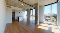 This single-floor unit showcases an industrial-modern aesthetic defined by exposed concrete ceilings with visible pipe details, raw concrete columns, and large floor-to-ceiling windows that flood the interior with natural light and frame expansive urban views, including a notable arch bridge on the horizon.

The open-concept living and dining area features warm hardwood flooring and connects seamlessly to a well-appointed kitchen. The kitchen is equipped with white cabinetry, stainless steel appliances including a refrigerator, microwave, and range, a dark tile backsplash, and a central island with a gray quartz countertop and undermount sink. A pendant light anchors the kitchen space, complementing the industrial character of the exposed ceiling above.

Two bedrooms offer a quieter retreat, both carpeted in deep navy blue and accented by the same exposed concrete column detail found throughout the unit. Each room benefits from window access and ceiling fan fixtures. The bathroom is clean and functional, featuring a white subway-tiled tub-shower combo, a white vanity with integrated sink, and teal vinyl flooring that adds a distinct pop of color to the otherwise neutral palette.