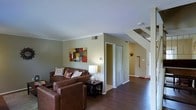 This two-story residence offers a well-organized layout designed for comfortable everyday living. The main level is anchored by a spacious living area that immediately draws attention with its rich dark hardwood flooring, which adds warmth and depth to the space. Crown molding along the ceiling provides a refined architectural detail, while the textured ceiling adds a classic touch overhead. The walls are finished in a neutral olive-green tone, creating a welcoming and cohesive atmosphere throughout the main floor. A staircase is prominently positioned, connecting the two levels and serving as a functional architectural element within the open layout. Natural light filters into the entry area through a window near the front door, brightening the space and highlighting the clean white trim and baseboards. A louvered closet door near the living area provides convenient storage access. The living room itself is generously sized, accommodating substantial seating with ease. Vaulted or angled ceiling lines near the staircase add visual interest and a sense of height to the interior. Overall, the space balances practical design with understated aesthetic details, offering a functional and inviting environment across both floors.