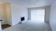 This single-floor space offers a clean, functional layout designed for comfortable everyday living. The main living area is a standout feature, showcasing a classic fireplace with a tiled hearth and surround that serves as a natural focal point within the room. Plush wall-to-wall carpeting in a neutral tone runs throughout, providing a soft and inviting underfoot experience that complements the bright, airy atmosphere.

A full-width window wall dressed with vertical blinds dominates one side of the living area, allowing an abundance of natural light to pour in and creating a sense of openness and spaciousness. The white walls are clean and unadorned, offering a versatile blank canvas that suits a wide range of interior styles and furnishing choices.

The textured ceiling adds a subtle architectural detail consistent with the overall aesthetic of the space. The combination of the generous window coverage, neutral color palette, and carpeted flooring gives the living area a warm yet understated character. Overall, this space presents a straightforward and well-maintained environment with key comfort-oriented features, including its charming fireplace and light-filled living room, making it a welcoming and adaptable home.