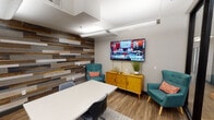 This well-appointed single-floor office space blends contemporary design with warm, inviting elements to create a functional and visually engaging work environment. The centerpiece of the space is a multipurpose conference and lounge room, defined by a striking floor-to-ceiling reclaimed wood accent wall composed of varied planks in rich tones of brown, gray, and white, lending texture and character to the interior. Wood-look flooring runs throughout, complementing the overall aesthetic. The room is furnished with a clean white conference table and modern task chairs, alongside a pair of teal mid-century-style armchairs accented with coral throw pillows, creating an inviting lounge area. A warm-toned yellow credenza serves as both a storage and display surface beneath a large wall-mounted flat-screen TV, ideal for presentations or collaborative viewing. Dual glass whiteboard panels on an adjacent wall provide a practical surface for notes and brainstorming sessions. The space is enclosed by sleek floor-to-ceiling black-framed glass partitions, offering visual openness while maintaining separation from the outer workstation area, which features desktop computer setups. Overhead linear pendant lighting provides bright, even illumination throughout. The overall ambiance is modern, clean, and professionally designed to support both focused work and collaborative interaction.