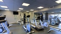This fitness center is designed to support a comprehensive workout experience within a compact, efficiently organized layout. The space features a dark, durable flooring surface well-suited for high-traffic exercise use, paired with neutral gray walls that contribute to a clean, focused atmosphere. Overhead, a drop ceiling with large flush-panel light fixtures provides bright, even illumination throughout the room. A full-length mirrored wall along one side visually expands the space while allowing users to monitor their form during workouts. The equipment selection is well-rounded, including a multi-function cable and weight machine for strength training, an elliptical cross-trainer, a treadmill, and additional cardio equipment. An instructional exercise poster is mounted near the strength training station, offering guidance for users of all fitness levels. A wall-mounted television is positioned for easy viewing from the cardio equipment area. The overall design prioritizes functionality and practicality, making this an accessible and well-appointed fitness facility for daily use.