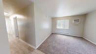 Located at 4415 Shepard Rd NE in Albuquerque, New Mexico, this single-floor unit offers three bedrooms and one bathroom within a compact, efficiently designed layout. The interior features a neutral color palette of soft beige and white tones throughout, creating a clean and consistent aesthetic across all rooms. Flooring transitions between plush carpeting in the bedrooms and living areas, and hardwood-style planks near the entryway and transitional spaces. Each bedroom is equipped with a sliding or hinged closet featuring built-in shelving, offering practical storage solutions. The spacious primary bedroom includes a wall-mounted air conditioning unit and a baseboard heater beneath a large window dressed with vertical blinds, providing both natural light and climate control. Additional bedrooms maintain the same carpeted finish and neutral walls. The bathroom features a white drop-in sink with brushed nickel faucet, a mirrored medicine cabinet, and tile surround near the tub area. The entry area includes a coat closet and opens into the main living space. Textured ceilings are present throughout, adding subtle architectural character to the otherwise straightforward and functional design of this Albuquerque residence.