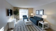 This single-floor space centers around a well-appointed living room that balances comfort and style. A large sliding glass door serves as the room's focal point, flooding the space with natural light and offering a view to the outside. Neutral beige walls and a textured ceiling create a clean, understated backdrop throughout.

The living area is generously sized, accommodating a tufted navy blue sofa paired with a matching accent chair, both positioned around a white coffee table atop a bold geometric-patterned area rug. The soft carpeting underfoot adds warmth and texture to the room. A wall-mounted television anchors one side of the space, while white side tables with lamps provide functional lighting and symmetry.

A large potted fiddle-leaf fig tree adds a natural, organic element to the corner of the room, complementing the greenery visible through the sliding door. Framed artwork on the walls contributes a polished, finished look. The overall ambiance is bright, clean, and inviting, with a cohesive design that emphasizes casual comfort and everyday livability.
