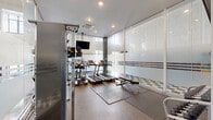 Located at 50 West 4th Street in downtown Winston-Salem, NC, this well-appointed fitness center offers a comprehensive workout experience within a compact, efficiently designed space. The exercise room is enclosed by floor-to-ceiling frosted glass panel walls with horizontal stripe detailing, lending the space a modern, sleek aesthetic while allowing diffused natural light to filter through from surrounding windows positioned near the ceiling line.

The room is equipped with multiple treadmills and an elliptical trainer, all positioned to face a wall-mounted flat-screen television for entertainment during cardio sessions. A multi-function cable machine provides options for a wide range of strength training exercises, complemented by a rack of dumbbells in varying weights and an adjustable workout bench for free weight training. Stability balls round out the strength and conditioning offerings.

Flooring transitions thoughtfully throughout the space, with rubber matting in the main workout areas providing cushioning and grip, while a checkered tile floor defines a secondary zone within the glass-enclosed section. Recessed ceiling lighting ensures bright, even illumination throughout. A ceiling-mounted HVAC unit maintains climate control, ensuring a comfortable environment year-round. The overall design is clean, functional, and modern, making this fitness center a practical amenity for daily use.