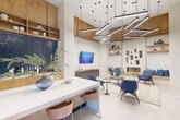 Located at 352 S Foster Dr in Baton Rouge, Louisiana, this thoughtfully designed single-floor space offers an open, airy layout that feels expansive thanks to its dramatic soaring ceilings and clean white walls. Large-format light tile flooring runs throughout, lending a cohesive and polished aesthetic to the entire space.

The layout is organized into several distinct functional zones. A comfortable lounge area anchors one end of the space, featuring a built-in linear fireplace framed by decorative wall niches and open wood shelving displaying curated décor. Blue upholstered chairs and a neutral area rug create an inviting seating arrangement, complemented by a wall-mounted flat-screen television and a sleek media credenza.

A bar-height counter with a white stone surface and stylish seating serves as a social gathering point, backed by rich blue tile and warm wood cabinetry. A standalone open wood shelving unit provides both storage and visual interest nearby. A dedicated workspace or meeting area is visible toward the rear, fitted with work surfaces and seating for focused use.

Overhead, an eye-catching geometric arrangement of suspended linear pendant lights adds a bold, contemporary design statement. Elevated warm-toned wood cabinetry lines the upper walls throughout, reinforcing the space's cohesive material palette. Natural light filters in through windows, while recessed lighting ensures the space remains well-lit throughout.