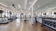 Located at 1920 Exchange Dr in Greenville, NC, this single-floor fitness and amenity facility offers a comprehensive and thoughtfully designed space for exercise and relaxation. The expansive main gym area is flooded with natural light through large black-framed windows and features durable tile flooring, ceiling fans, and recessed lighting accented by subtle pink ambient illumination. The fitness floor is outfitted with a wide array of equipment, including treadmills, ellipticals, stair climbers, strength machines, and free weights with a dedicated weight bench area backed by full-length mirrors. Textured mosaic accent walls add visual interest throughout the gym. Adjacent to the workout floor is a stylish lounge area with modular seating, colorful accent pillows, a distinctive patterned ceiling, and curated wall art, creating an inviting space to unwind. A private meeting room enclosed by glass doors features a round table, modern rolling chairs in teal, patterned carpet, and tall windows offering exterior views. A dedicated corridor leads to restrooms and includes a water fountain station, finished with deep teal walls and patterned tile flooring for a polished, cohesive aesthetic throughout the facility.