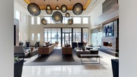 This elegantly appointed club room offers an impressive gathering space defined by its soaring double-height ceilings and an eye-catching installation of large, sculptural globe pendant lights in an intricate lattice design. A warm wood-paneled ceiling accent adds a natural contrast to the crisp white walls, while floor-to-ceiling glass doors and clerestory windows flood the interior with abundant natural light and frame sweeping views of the waterfront and city skyline beyond.

The room is thoughtfully arranged with multiple seating areas, featuring plush sofas and accent chairs in warm caramel and charcoal tones set atop patterned area rugs. A sleek fireplace anchors one side of the room, providing a focal point that enhances the space's inviting ambiance. The polished tile flooring near the entry transitions seamlessly into the carpeted lounge areas, guiding movement throughout the room.

A glass-enclosed area along one side adds architectural interest and a sense of privacy, while the open layout encourages both intimate conversation and larger social gatherings. The overall design balances contemporary sophistication with a welcoming warmth, making this club room an exceptional amenity space for residents and guests alike.