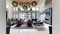 This elegantly appointed club room offers an impressive gathering space defined by its soaring double-height ceilings and an eye-catching installation of large, sculptural globe pendant lights in an intricate lattice design. A warm wood-paneled ceiling accent adds a natural contrast to the crisp white walls, while floor-to-ceiling glass doors and clerestory windows flood the interior with abundant natural light and frame sweeping views of the waterfront and city skyline beyond.

The room is thoughtfully arranged with multiple seating areas, featuring plush sofas and accent chairs in warm caramel and charcoal tones set atop patterned area rugs. A sleek fireplace anchors one side of the room, providing a focal point that enhances the space's inviting ambiance. The polished tile flooring near the entry transitions seamlessly into the carpeted lounge areas, guiding movement throughout the room.

A glass-enclosed area along one side adds architectural interest and a sense of privacy, while the open layout encourages both intimate conversation and larger social gatherings. The overall design balances contemporary sophistication with a welcoming warmth, making this club room an exceptional amenity space for residents and guests alike.