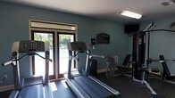 This fitness center offers a well-appointed workout environment designed to meet a range of exercise needs. The space is finished with cool-toned, teal-painted walls and a neutral ceiling, creating a clean and focused atmosphere conducive to exercise. Large glass double doors serve as a prominent architectural feature, flooding the interior with natural light while providing a direct view of the outdoor pool area beyond — a refreshing visual backdrop during workouts. The flooring is covered with dark gym-appropriate matting, offering comfort and durability underfoot. The facility is equipped with essential cardio machines, including treadmills and an elliptical trainer, all positioned to take advantage of the natural light and outdoor views. A strength training station is also present, providing options for resistance-based exercise. A posted fitness center rules sign on the wall indicates a managed and maintained facility. An overhead fluorescent light fixture supplements the natural light, ensuring the space remains well-lit at all times. A wall-mounted television adds an entertainment option for users during their workout sessions. Overall, this fitness center delivers a practical, comfortable, and light-filled environment suitable for a variety of fitness routines.