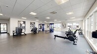 This single-floor fitness center at 375 E Main St in Branford, CT is a comprehensively equipped facility designed to support a full range of workout routines. The space features a clean, modern aesthetic with white walls, a drop tile ceiling with recessed panel lighting, and durable dark rubber flooring throughout. Large windows along multiple walls allow abundant natural light to fill the interior while offering views of the surrounding neighborhood, creating an open and energizing atmosphere. The layout is spacious and well-organized, with distinct zones dedicated to different types of training. The cardio section is outfitted with a generous row of treadmills, elliptical machines, and rowing ergometers — all arranged to maximize floor space and user comfort. A dedicated free weights area includes a full dumbbell rack with adjustable benches and a spin bike. The strength training zone features blue cable machines and multi-functional weight stations, complemented by decorative geometric wall art that adds visual character to the space. Additional equipment includes medicine balls, stability balls, and kettle bells neatly stored on dedicated racks. Full-length mirrors along one wall enhance the sense of depth and allow users to monitor their form. The facility is climate-controlled with ceiling-mounted HVAC units, ensuring year-round comfort.