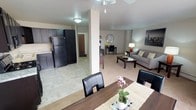 This single-floor residence is designed around an open-concept layout that seamlessly connects the kitchen, dining, and living areas into one cohesive space. The kitchen is outfitted with dark cabinetry and black appliances, including a full-size refrigerator, and features light tile flooring that extends through the entry and kitchen zone. A structural column subtly defines the boundary between the kitchen and the central dining area, where a dark wood dining table with black upholstered chairs provides a functional and stylish gathering space. Beyond the dining area, plush carpeting begins and flows throughout the living room, which is furnished with a neutral sofa, a rectangular coffee table, and warm accent lighting. A decorative wall mirror and framed artwork add personality to the walls. A secondary desk or workstation area is visible deeper in the space, offering a dedicated spot for work or study. The unit is bathed in ambient artificial lighting with a ceiling fan overhead, and the entryway with its solid door is visible in the background, completing the practical and comfortable layout of this well-organized living space.