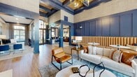 Located at 13625 Haven Ridge Ln in Charlotte, NC, this beautifully appointed single-story clubhouse exemplifies upscale community living through its refined architectural design and carefully curated interiors. The main lounge area commands attention with soaring coffered ceilings accented by dark navy beams and a striking gold chandelier, creating an atmosphere of understated luxury. Rich navy blue panel millwork lines the walls throughout, complemented by warm wood-look flooring that adds a welcoming, residential feel.

Seating areas are thoughtfully arranged to encourage both socializing and relaxation, featuring a mix of tufted caramel leather banquette seating, plush neutral sofas, and accent chairs layered over patterned area rugs. A classic brick fireplace topped with a wall-mounted television anchors one end of the lounge, providing a cozy focal point.

Floor-to-ceiling windows and elegant French doors flood the interior with natural light, framed by dramatic floor-length floral draperies that add a bold, artistic touch. A dedicated meeting or dining nook features a round table surrounded by velvet upholstered chairs, ideal for small gatherings. Adjacent spaces visible through glass-paneled doors suggest additional work or amenity areas. The overall ambiance is sophisticated yet inviting, blending contemporary design with warm, clubhouse-style comfort.