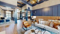 Located at 13625 Haven Ridge Ln in Charlotte, NC, this beautifully appointed single-story clubhouse exemplifies upscale community living through its refined architectural design and carefully curated interiors. The main lounge area commands attention with soaring coffered ceilings accented by dark navy beams and a striking gold chandelier, creating an atmosphere of understated luxury. Rich navy blue panel millwork lines the walls throughout, complemented by warm wood-look flooring that adds a welcoming, residential feel.

Seating areas are thoughtfully arranged to encourage both socializing and relaxation, featuring a mix of tufted caramel leather banquette seating, plush neutral sofas, and accent chairs layered over patterned area rugs. A classic brick fireplace topped with a wall-mounted television anchors one end of the lounge, providing a cozy focal point.

Floor-to-ceiling windows and elegant French doors flood the interior with natural light, framed by dramatic floor-length floral draperies that add a bold, artistic touch. A dedicated meeting or dining nook features a round table surrounded by velvet upholstered chairs, ideal for small gatherings. Adjacent spaces visible through glass-paneled doors suggest additional work or amenity areas. The overall ambiance is sophisticated yet inviting, blending contemporary design with warm, clubhouse-style comfort.