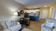 This single-floor space showcases an open-concept layout that seamlessly connects the kitchen, dining, and living areas, creating a functional and inviting environment. The kitchen is a standout feature, equipped with white shaker-style cabinetry, a stainless steel refrigerator, and a matching stainless steel range with microwave above. A tiled backsplash adds texture, while pendant lighting suspended above the kitchen island provides both task lighting and visual interest. The island itself features a light stone countertop with a rich blue base, offering bar-style seating with dark round stools — ideal for casual dining or entertaining. The dining area accommodates a dark-finished table with bench seating, positioned conveniently between the kitchen and living space. The living area is furnished with comfortable upholstered seating and complemented by a floor lamp that adds warm ambient light. Warm-toned luxury vinyl plank flooring runs throughout, tying all areas together cohesively. Neutral beige walls and recessed lighting contribute to the bright, welcoming atmosphere of this thoughtfully arranged single-level space.