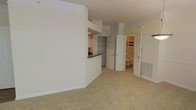 This single-floor residence offers a clean, functional layout designed for comfortable everyday living. The main living area is finished with plush wall-to-wall carpeting and painted in neutral tones throughout, creating a bright and welcoming atmosphere. Crown molding adds a subtle architectural detail that frames the space with a classic touch.

The kitchen connects to the living area via a pass-through counter with a dark-toned surface, providing a convenient open connection between the two spaces while maintaining a defined separation. Cabinetry is visible in the kitchen area, offering ample storage. A built-in shelving unit adjacent to the kitchen provides additional storage and display space.

Hallway access from the main living area leads to further rooms, including a full bathroom visible through an open door, finished with standard fixtures. A second door opens to additional private space, such as a bedroom or closet. A floor-level HVAC vent is integrated into the baseboard, ensuring efficient climate control throughout. White paneled doors are consistent across the unit, contributing to a cohesive and tidy interior aesthetic. The overall design is straightforward and well-maintained, making it a practical and move-in ready living space.