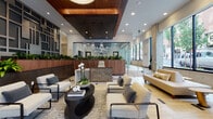 Located at 1425 SW Second Ave in Portland, Oregon, this beautifully designed lobby makes an immediate impression with its refined, contemporary aesthetic. The space centers around a striking walnut reception desk adorned with lush, integrated planters, complemented by suspended circular pendant lights that add a modern, sculptural quality to the overhead space. A coffered ceiling with rich wood paneling provides architectural depth and warmth, contrasting elegantly with the light-toned flooring accented by dark inlay details.

A standout feature is the large-scale geometric accent wall, composed of interlocking rectangular panels in layered grays and neutrals, lending the space a bold, urban character. Curated seating areas are thoughtfully arranged throughout, featuring contemporary lounge chairs and sofas in neutral tones, anchored by organic-shaped coffee tables that invite conversation and relaxation.

Floor-to-ceiling windows along the perimeter flood the interior with natural light while offering open views of the surrounding Portland streetscape. Carefully selected abstract artwork is displayed near the window-side seating alcove, adding a cultural touch to the welcoming atmosphere. The overall ambiance is polished and professional yet approachable, reflecting a high standard of design suited to its prominent downtown Portland address.